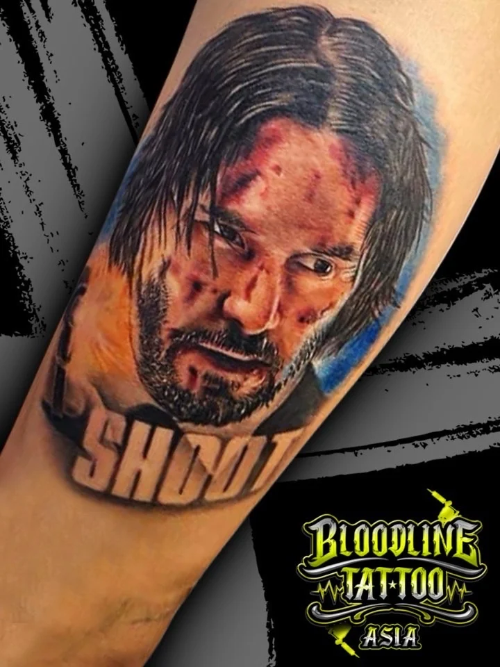 Colour portrait realism tattoo of John Wick with bruised face and bold SHOOT lettering below on the forearm at Bloodline Tattoo Bangkok.