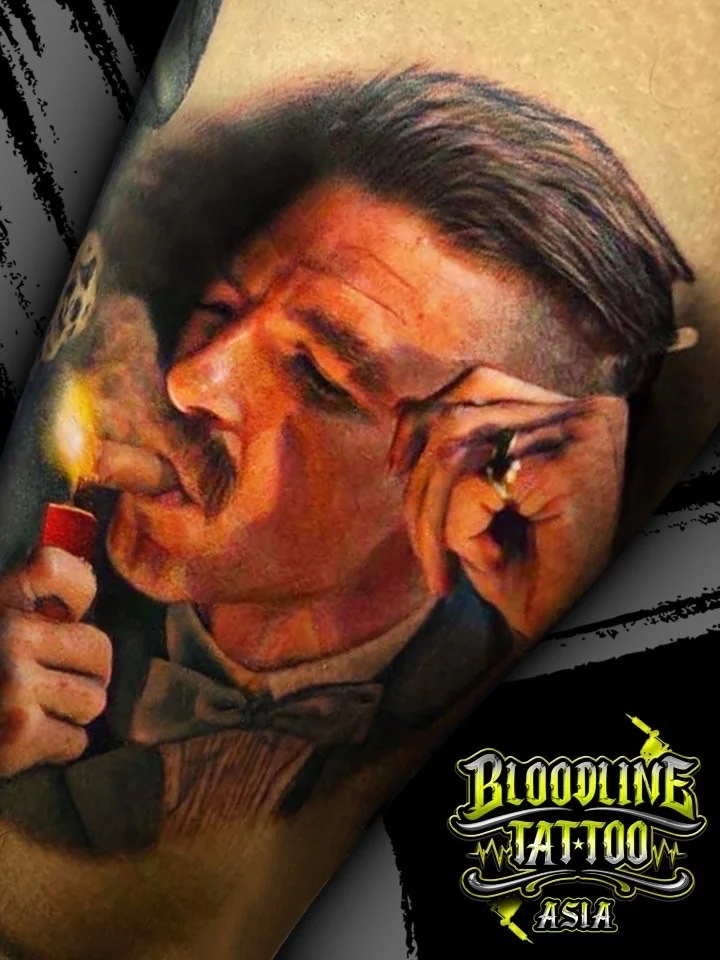 Colour portrait realism tattoo of Arthur Shelby lighting a cigar in a suit and bow tie at Bloodline Tattoo Bangkok.