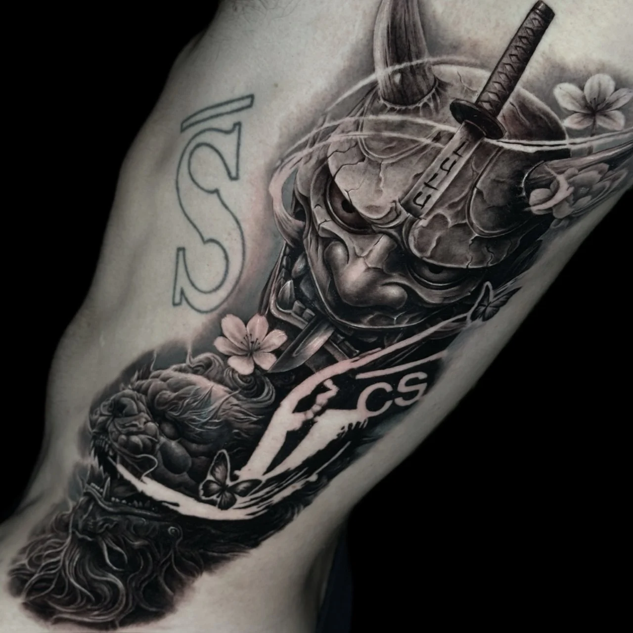 Black and grey Japanese torso tattoo of a stone Hannya mask pierced by a katana above a snarling Fu Dog with sakura at Bloodline Tattoo Bangkok.