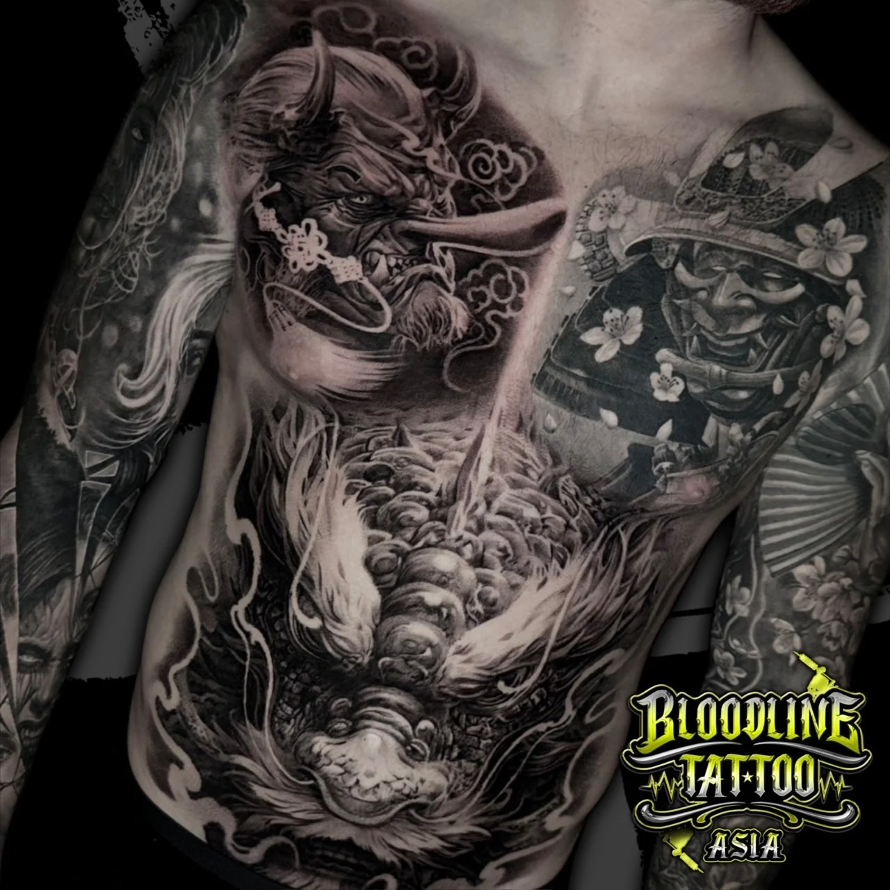 Black and grey Japanese chest and stomach tattoo with Tengu mask, samurai in kabuto, and upward dragon among clouds at Bloodline Tattoo Bangkok.