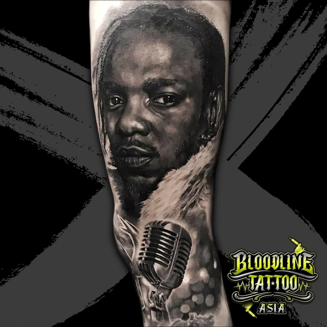 Black and grey portrait realism forearm tattoo of Kendrick Lamar with braids above a detailed vintage microphone at Bloodline Tattoo Bangkok.