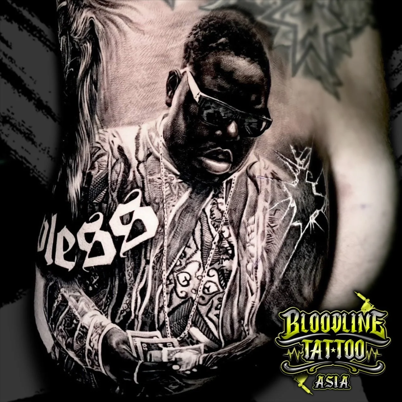 Black and grey portrait realism rib tattoo of The Notorious B.I.G. in sunglasses and Coogi sweater with Bless script and shattered glass effect at Bloodline Tattoo Bangkok.