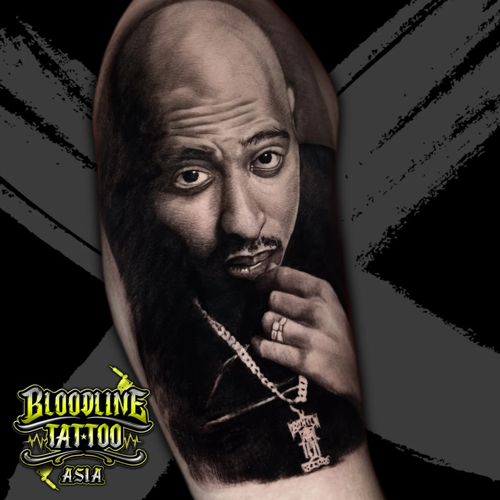 Black and grey portrait realism arm tattoo of Tupac Shakur with hand near mouth and a Death Row Records chain pendant at Bloodline Tattoo Bangkok.