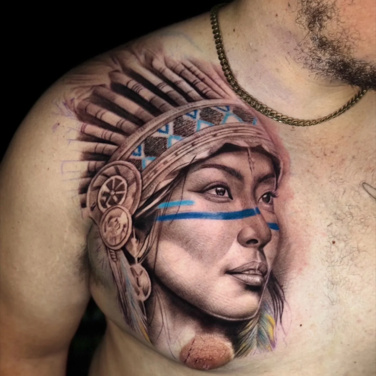A black and grey realism chest and shoulder portrait of an indigenous woman in a feathered headdress with face paint and patterned headband details, at Bloodline Tattoo Bangkok.