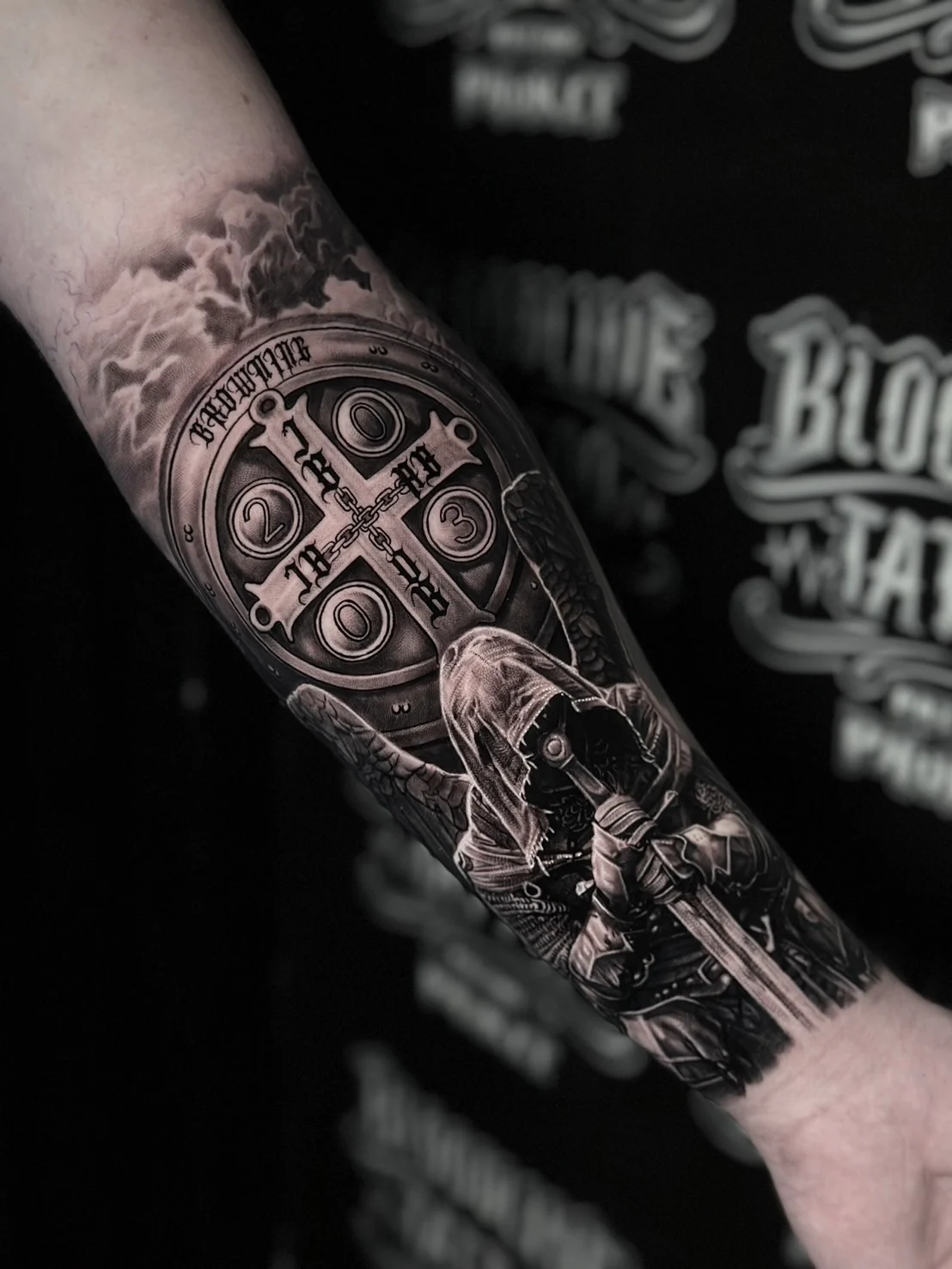 A black and grey realism forearm tattoo with Gothic "BROWNING" lettering above an ornate medallion and a hooded winged knight gripping a longsword, at Bloodline Tattoo Bangkok.